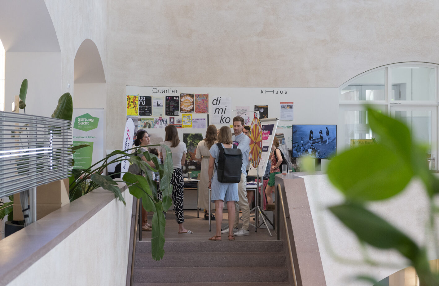 Einblick Basel K Haus Foto Basil Schubert Menschen stehen in einem hellen Raum mit Infostand und Postern.