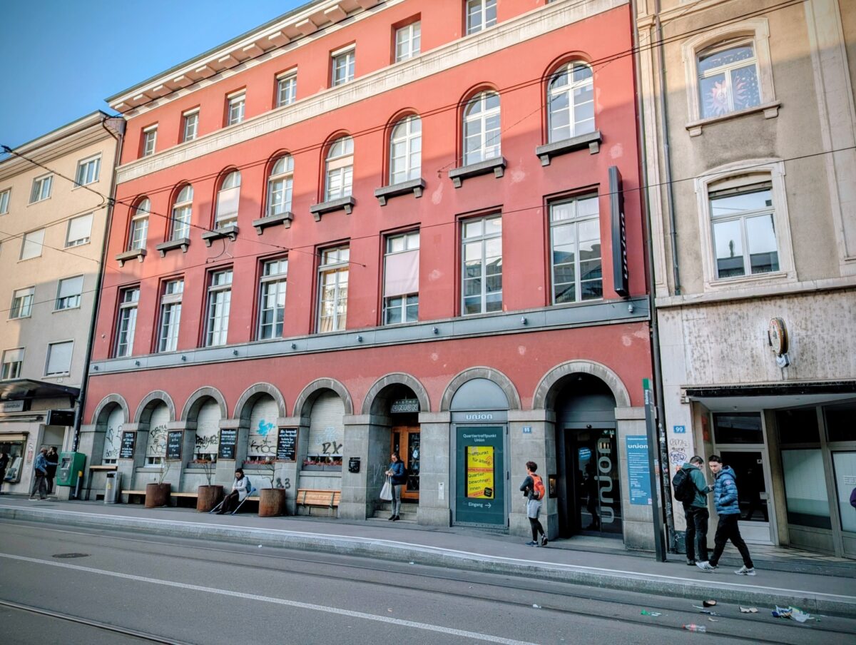 Ein rotes Gebäude mit Arkaden und Passanten auf einer Straße.