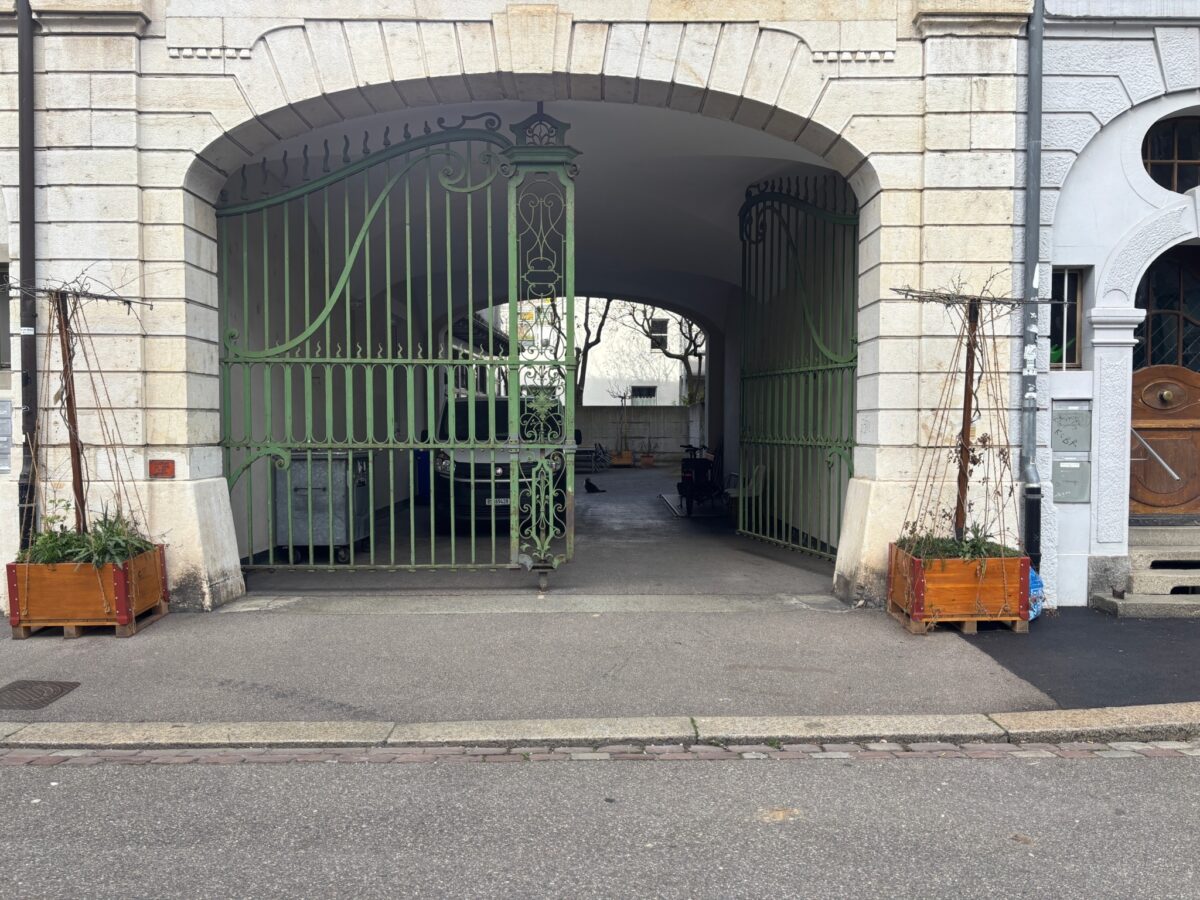 Ein grünes schmiedeeisernes Tor vor einem Steinbogen, flankiert von zwei Pflanzkisten.