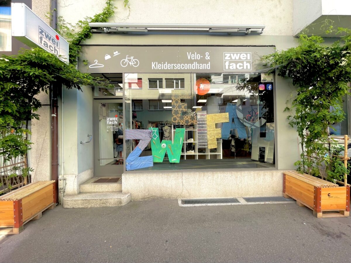 Ladenfront mit Schildern "Velo- & Kleidersecondhand" und "zweifach" sowie Pflanzen und Holzboxen davor.
