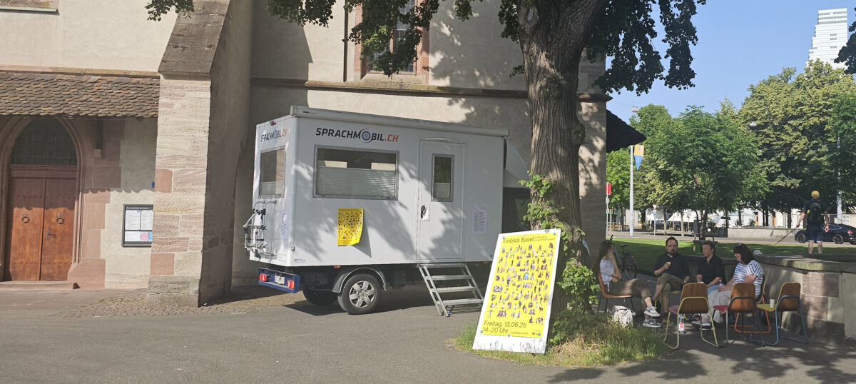 02 Einblick Ein weißer Sprachmobil-Wagen steht vor einem Gebäude mit Menschen auf Bänken daneben.