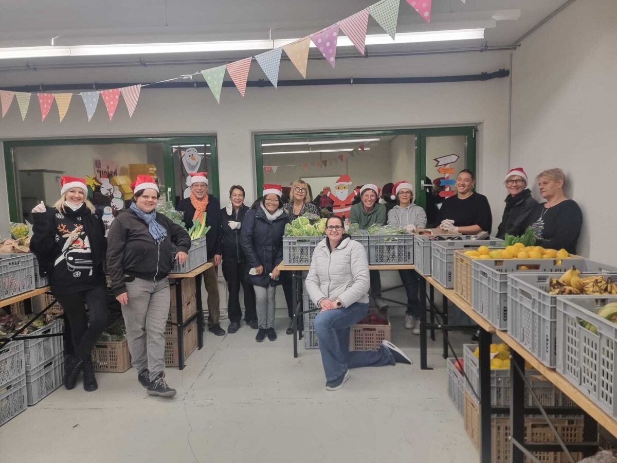 Eine Gruppe von Menschen steht in einem Raum mit Lebensmittelkisten und trägt Weihnachtshüte.