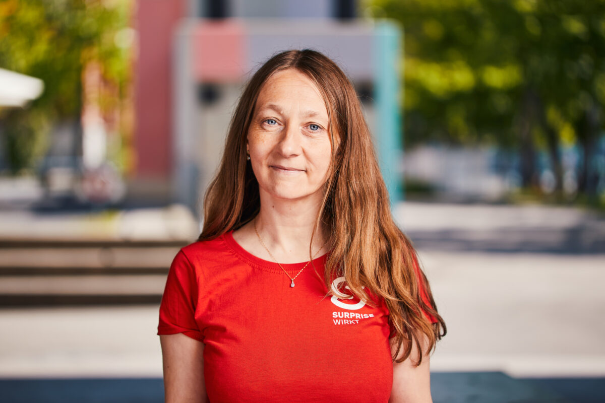 Danica Graf Portrait Lucia Hunziker Eine Frau mit langem Haar lächelt in einem roten T-Shirt mit Aufdruck.