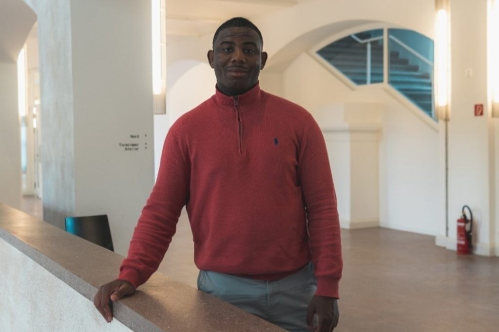 Gael Plo Baseljetzt Ein Mann in rotem Pullover lehnt an einem Geländer in einem hellen Gebäude.