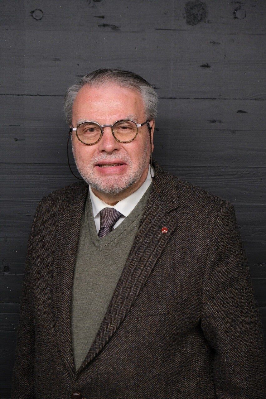 Marcel W Buess Ein älterer Mann mit Brille und Anzug steht vor einer dunklen Holzwand.