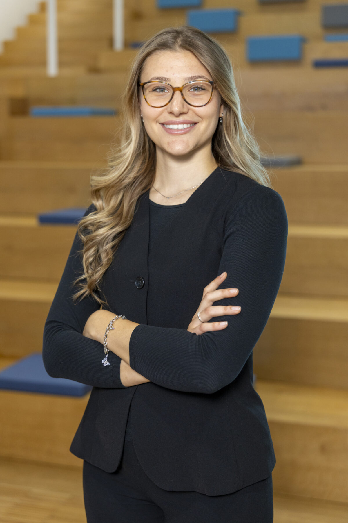 Sanja Radojicic Eine lächelnde Frau mit Brille steht mit verschränkten Armen in einem Raum mit Holzvertäfelung.