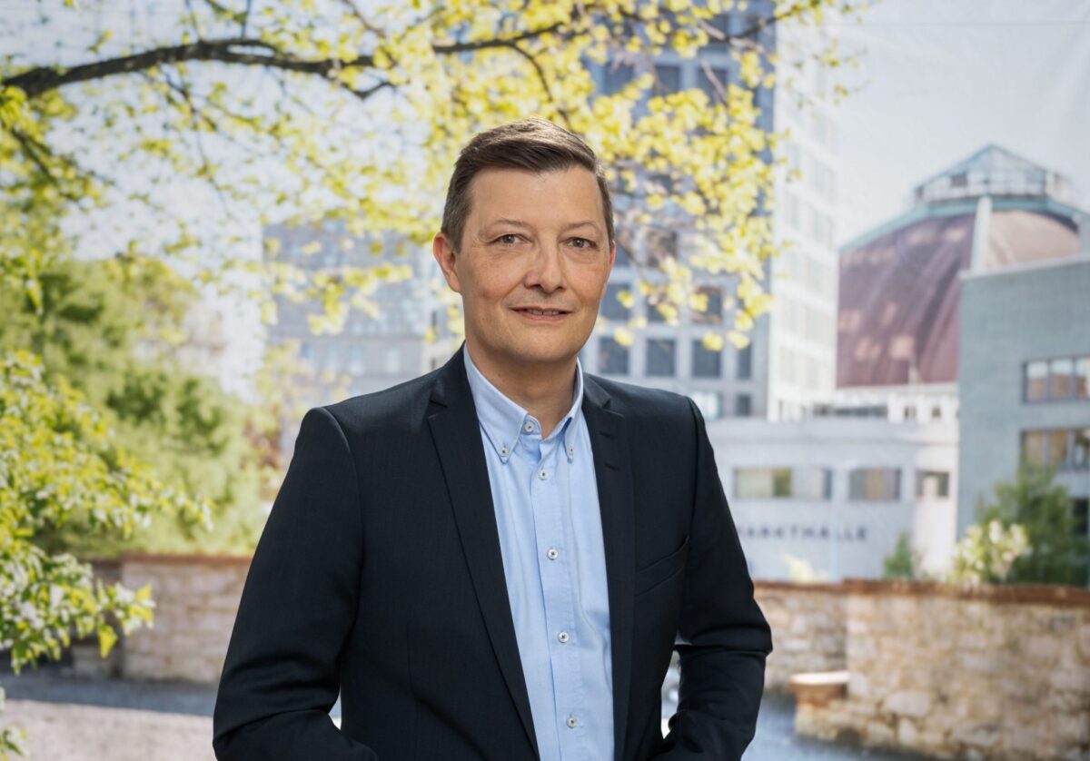 Ein Mann in Anzug steht vor einem Gebäude mit blühendem Baum.