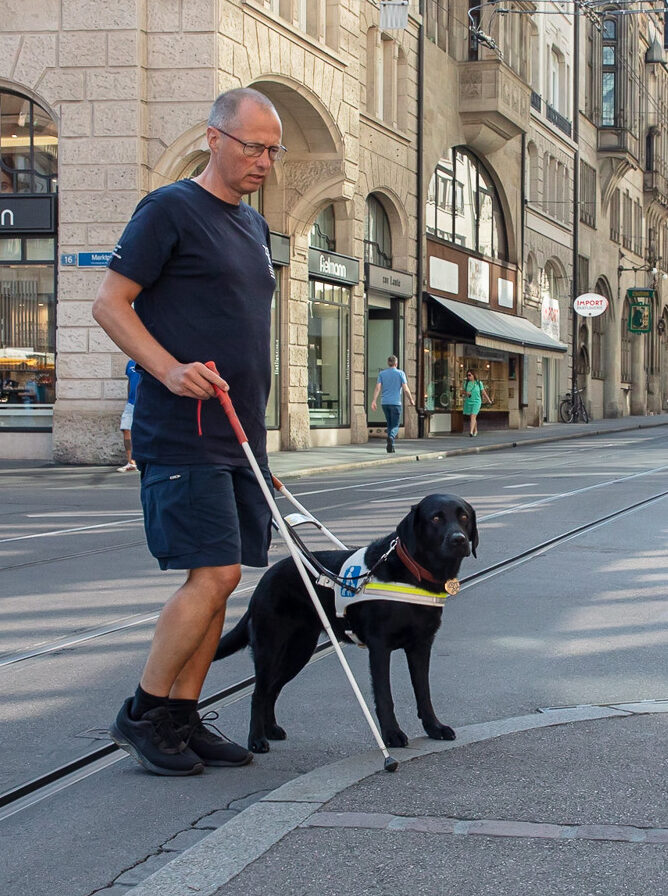 Blindenhundeschule Allschwil 20220816 BFHS RR Ein Mann mit einem Blindenhund überquert eine Straße in einer Stadt.
