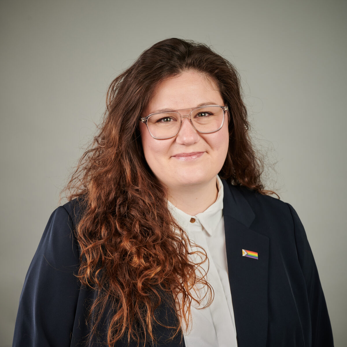 Jessica Brandenburger Eine Person mit Brille und Regenbogen-Anhänger lächelt in die Kamera.